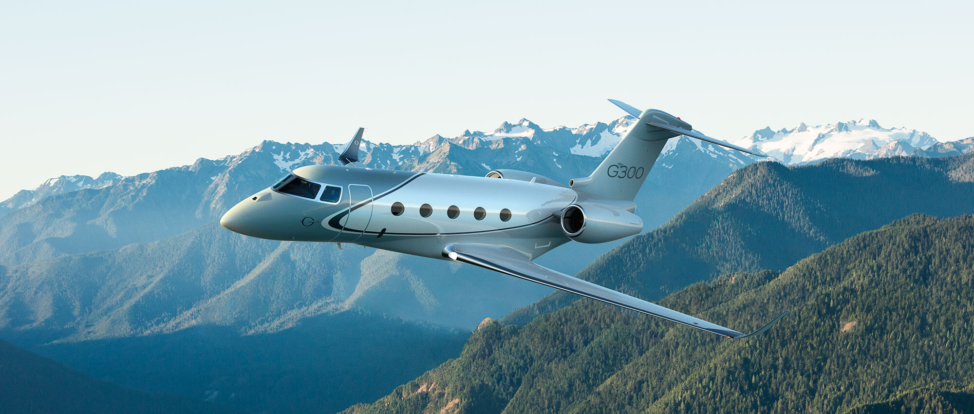 A Gulfstream G300 flies above forested mountains with snow-covered peaks in the distance.