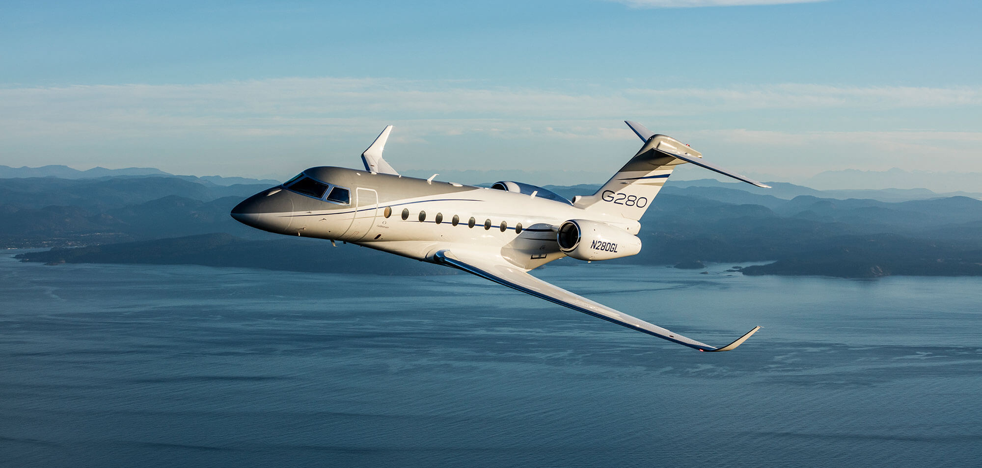 A Gulfstream G280 flies low over the blue water of an island-studded coastline.